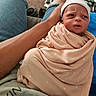 baby, infant, swaddled, blanket, hat, arm, denim_shorts, person, indoor, carpet, footwear, glass_table, curious_expression, skin, hand, couch, clothing, newborn, holding, wrapped