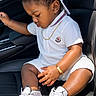 toddler, child, white_outfit, cap, sneakers, car_seat, gold_bracelet, gold_chain, black_hair, side_profile, cute, indoors, leather_seat, fashion, footwear, casual, portrait, young_child, sitting, daylight