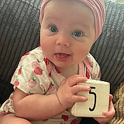 Maizie is registered to the contest to win money with this photo: baby, infant, headband, flower, wooden_block, number_five, couch, textured_cushion, pink_clothing, strawberry_pattern, cute, curious, tongue_out, sitting, indoors, natural_light, person, child, smiling, portrait