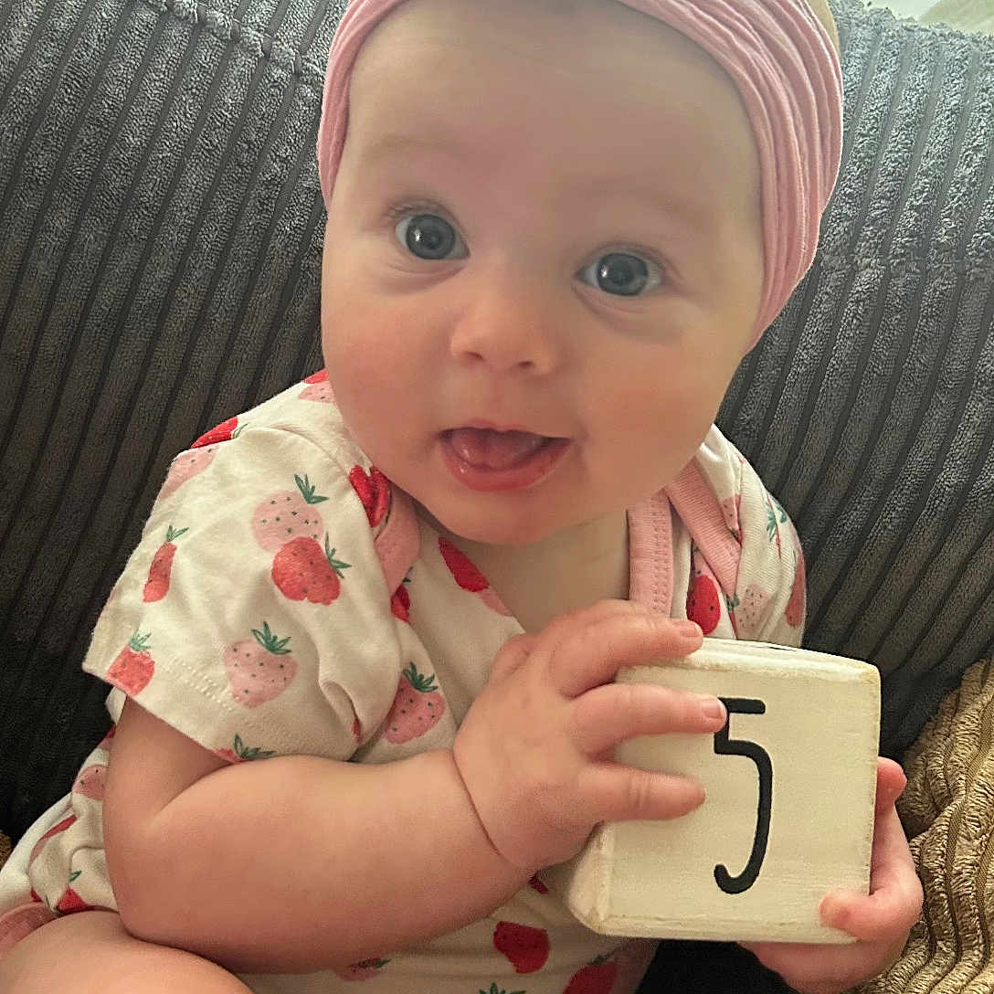 Maizie is registered to the contest to win money with this photo: baby, infant, headband, flower, wooden_block, number_five, couch, textured_cushion, pink_clothing, strawberry_pattern, cute, curious, tongue_out, sitting, indoors, natural_light, person, child, smiling, portrait