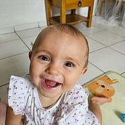 émilie a rejoint le concours — aidez-le/la à gagner de superbes lots ! baby, child, smiling, happy, crawling, dress, white_dress, patterned_clothing, floor, tile_floor, table, wooden_table, play_mat, cartoon_animal, indoor, home, person, face, head, eyes