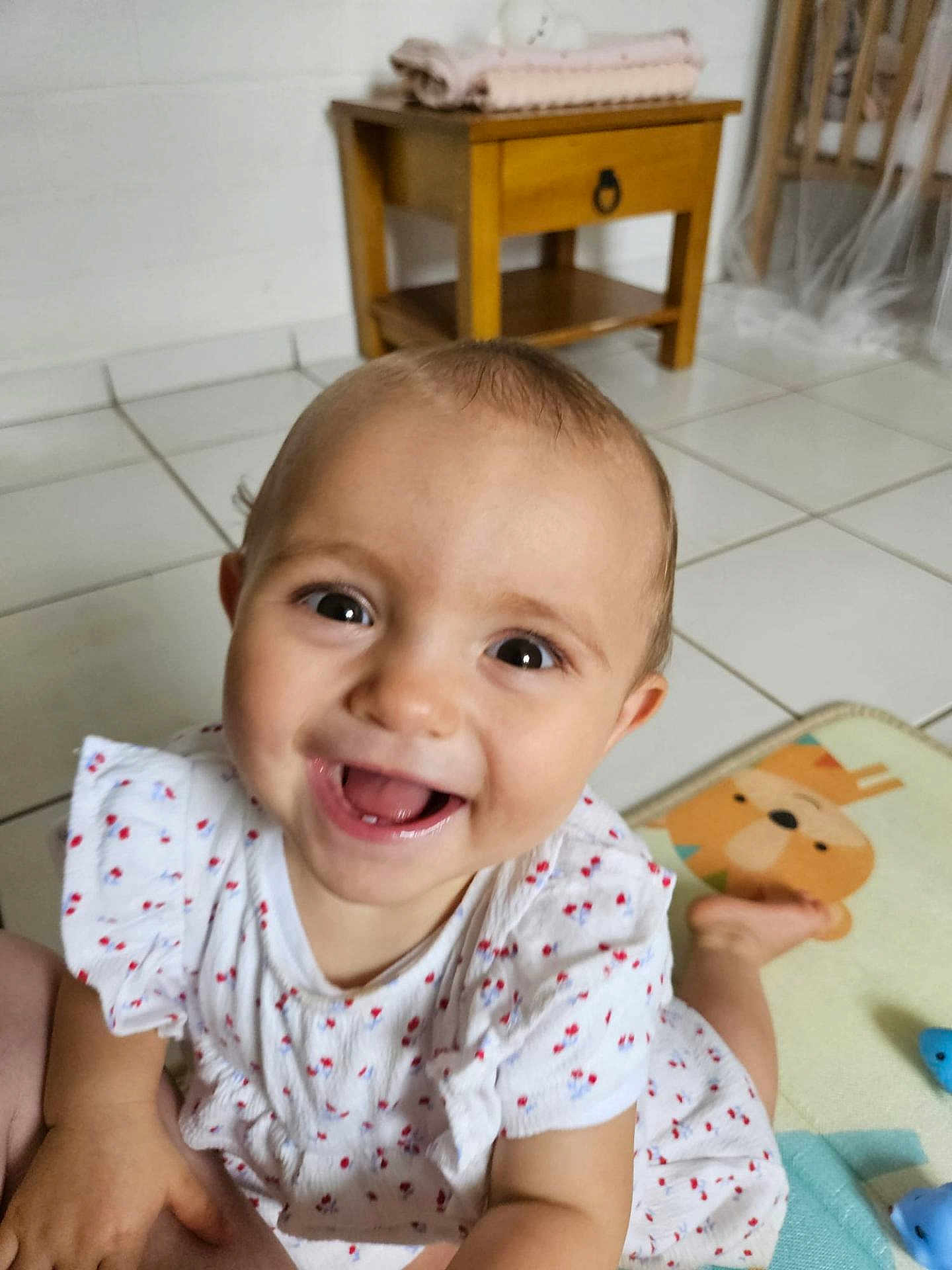 émilie a rejoint le concours — aidez-le/la à gagner de superbes lots ! baby, child, smiling, happy, crawling, dress, white_dress, patterned_clothing, floor, tile_floor, table, wooden_table, play_mat, cartoon_animal, indoor, home, person, face, head, eyes
