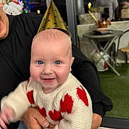 Sasha participe au concours pour gagner de l'argent avec cette photo : adult_hands, baby, blue_eyes, blurred_background, celebration, chair, christmas_tree, cozy, festive, green_carpet, happy, holding, indoor, joyful, party_hat, person, red_hearts, smiling, sweater, table