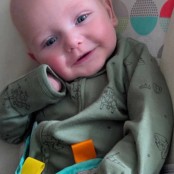 Ronin is registered to the contest to win money with this photo: baby, infant, smile, green_onesie, patterned_clothing, face, blue_eyes, seated, colorful_background, geometric_pattern, cozy, cute, child, portrait, happy, indoor, soft_lighting, young, relaxed, playful