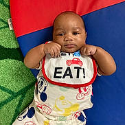 Grayson is registered to the contest to win money with this photo: baby, infant, bib, mickey_mouse, clothing, colorful, play_mat, green_carpet, red, blue, yellow, cute, smiling, child, person, portrait, lying_down, hands, raised_fists, indoor