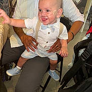Keniel is registered to the contest to win money with this photo: baby, child, infant, person, smiling, indoor, adult, clothing, bow_tie, suspenders, shoes, chair, table, floor, happy, medical_tube, nasal_cannula, hand, tongue, casual