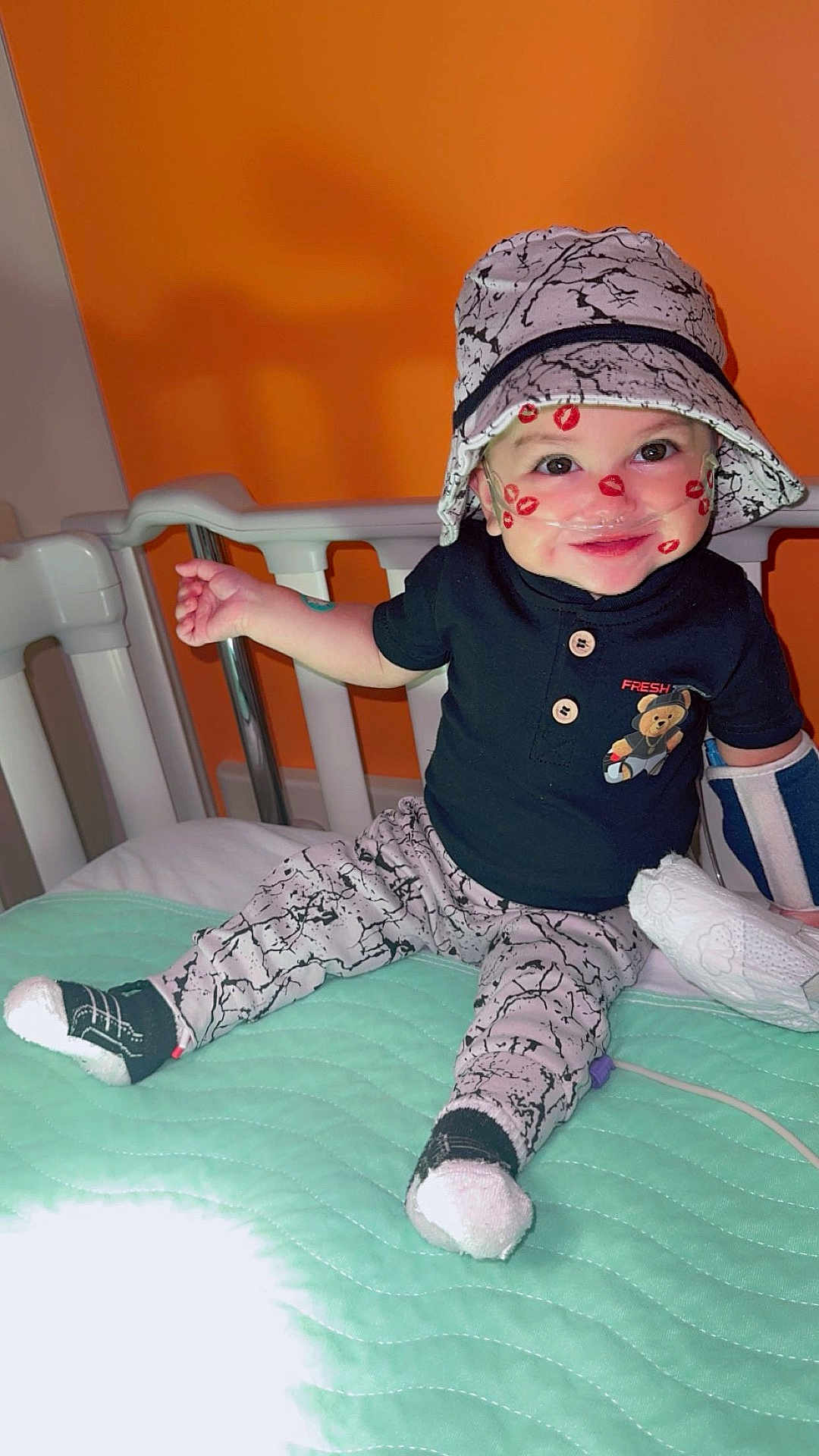 Keniel joined the competition — help win amazing prizes! toddler, child, hat, socks, hospital_bed, oxygen_tube, arm_brace, smile, face_paint, lipstick_marks, black_shirt, patterned_pants, blanket, medical_equipment, indoors, baby, cute, happy, person, sitting
