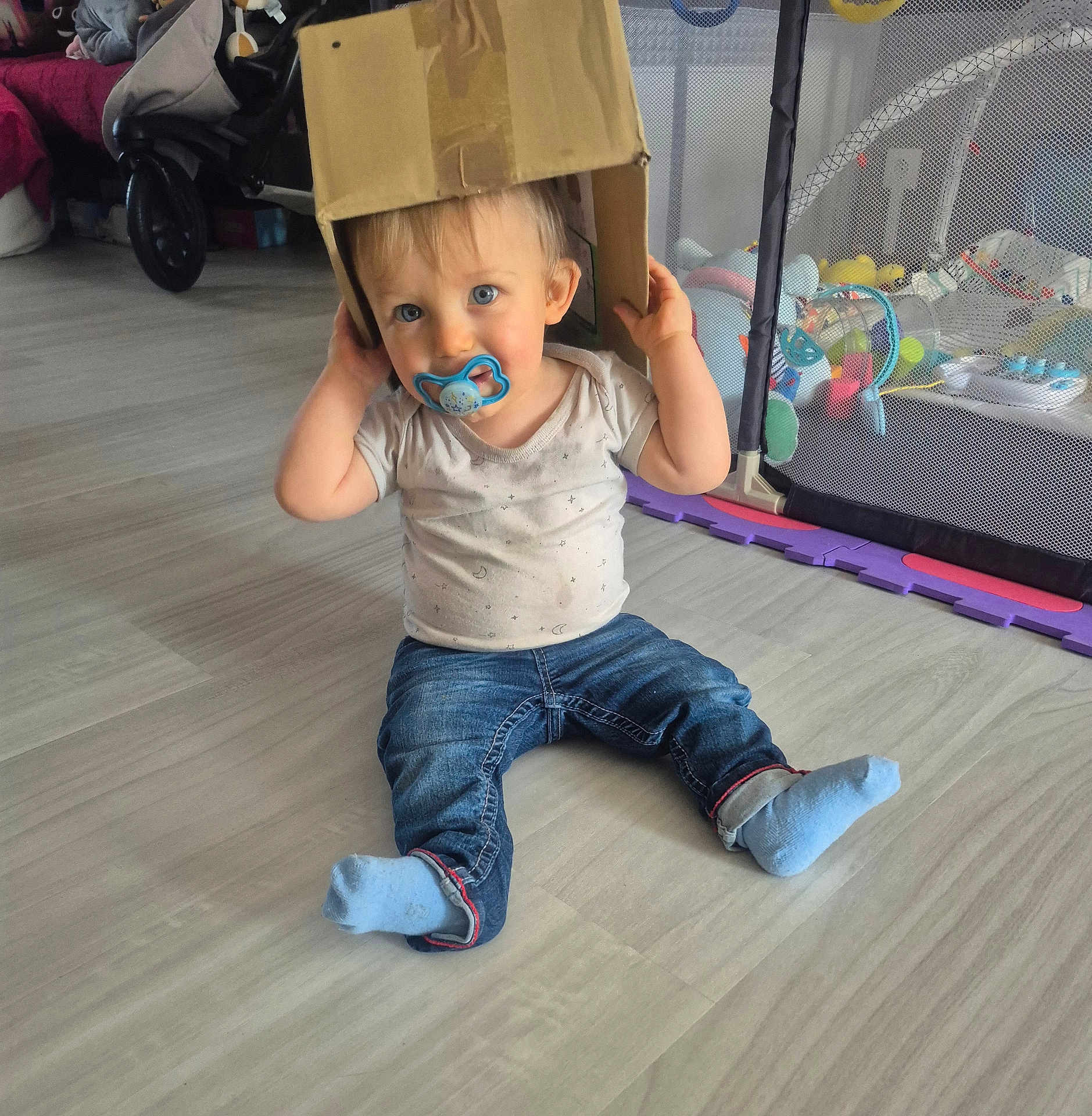 Gabriel participe au concours pour gagner de l'argent avec cette photo : toddler, child, pacifier, cardboard_box, jeans, socks, indoor, floor, toy, playpen, baby, person, cute, home, casual_clothing, sitting, blue_eyes, playful, infant, smiling