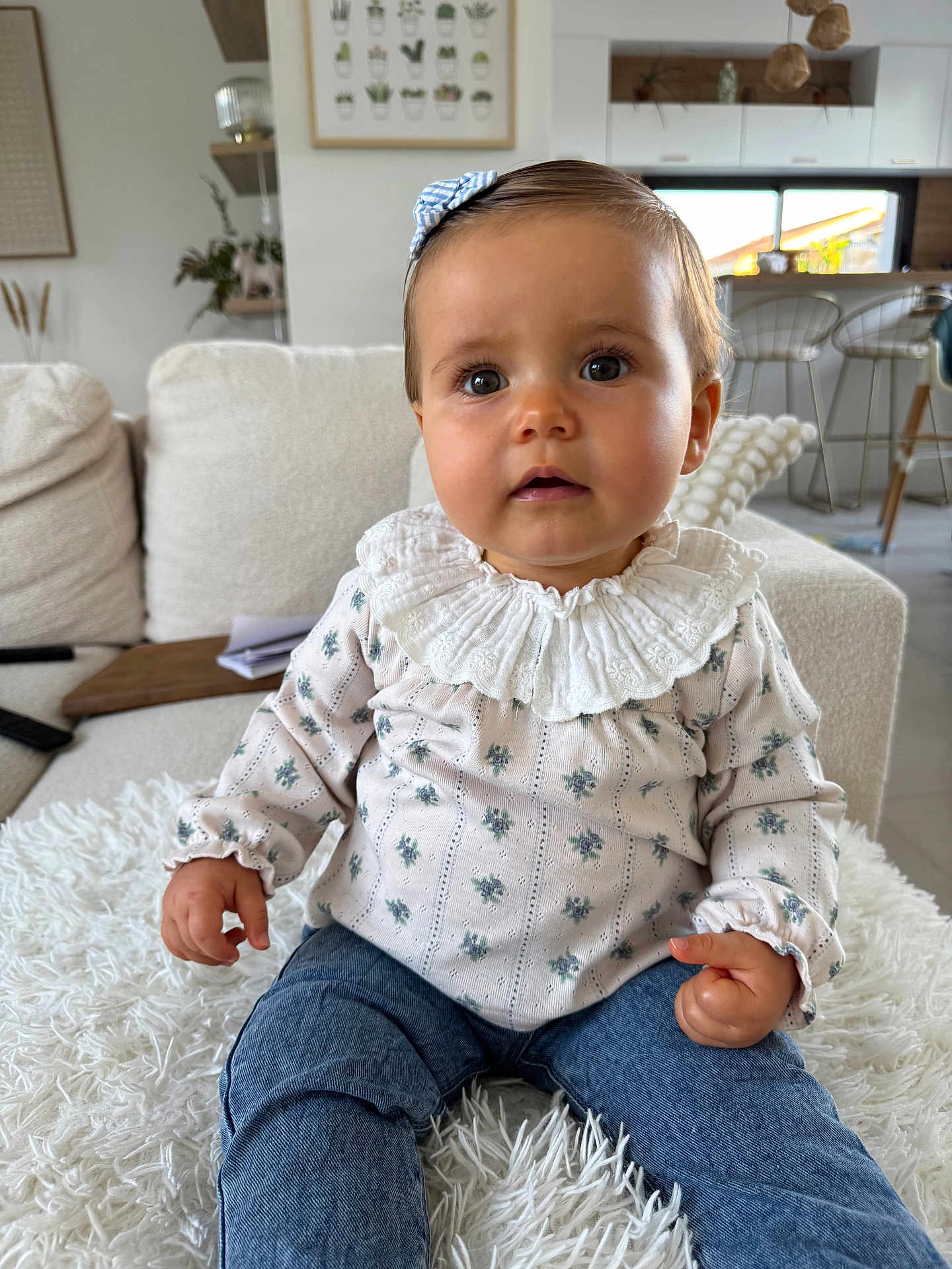 Julia participe au concours pour gagner de l'argent avec cette photo : baby, child, indoor, living_room, couch, rug, clothing, jeans, blouse, floral_pattern, curly_hair, face, hands, sitting, decor, plant, window, furniture, cute, portrait
