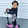 Giannis participe au concours pour gagner de l'argent avec cette photo : child, scooter, outdoor, pavement, nike, puma, sporty_clothing, black_jacket, gray_pants, toy, wheels, standing, young_child, curly_hair, white_wall, playtime, active, casual_wear, smiling, daylight