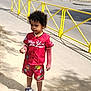 child, toddler, red_clothing, shorts, sneakers, curly_hair, sidewalk, yellow_fence, outdoor, daylight, street, holding_object, serious_expression, casual_wear, person, urban, shadow, tree, summer, walking