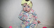 Brynlee is registered to the contest to win money with this photo: baby, blue_eyes, child, colorful, curious, cute, floral_pattern, headband, indoor, infant, neutral_background, onesie, pastel_colors, plastic_eggs, playtime, portrait, sitting, soft_texture, yellow, young_child