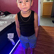 Ashton joined the competition — help win amazing prizes! child, boy, standing, black_tank_top, blue_shorts, sneakers, chain_necklace, bed, purple_light, wooden_floor, red_wall, indoor, furniture, cabinet, flooring, person, portrait, casual_clothing, hands_in_pockets, serious_expression