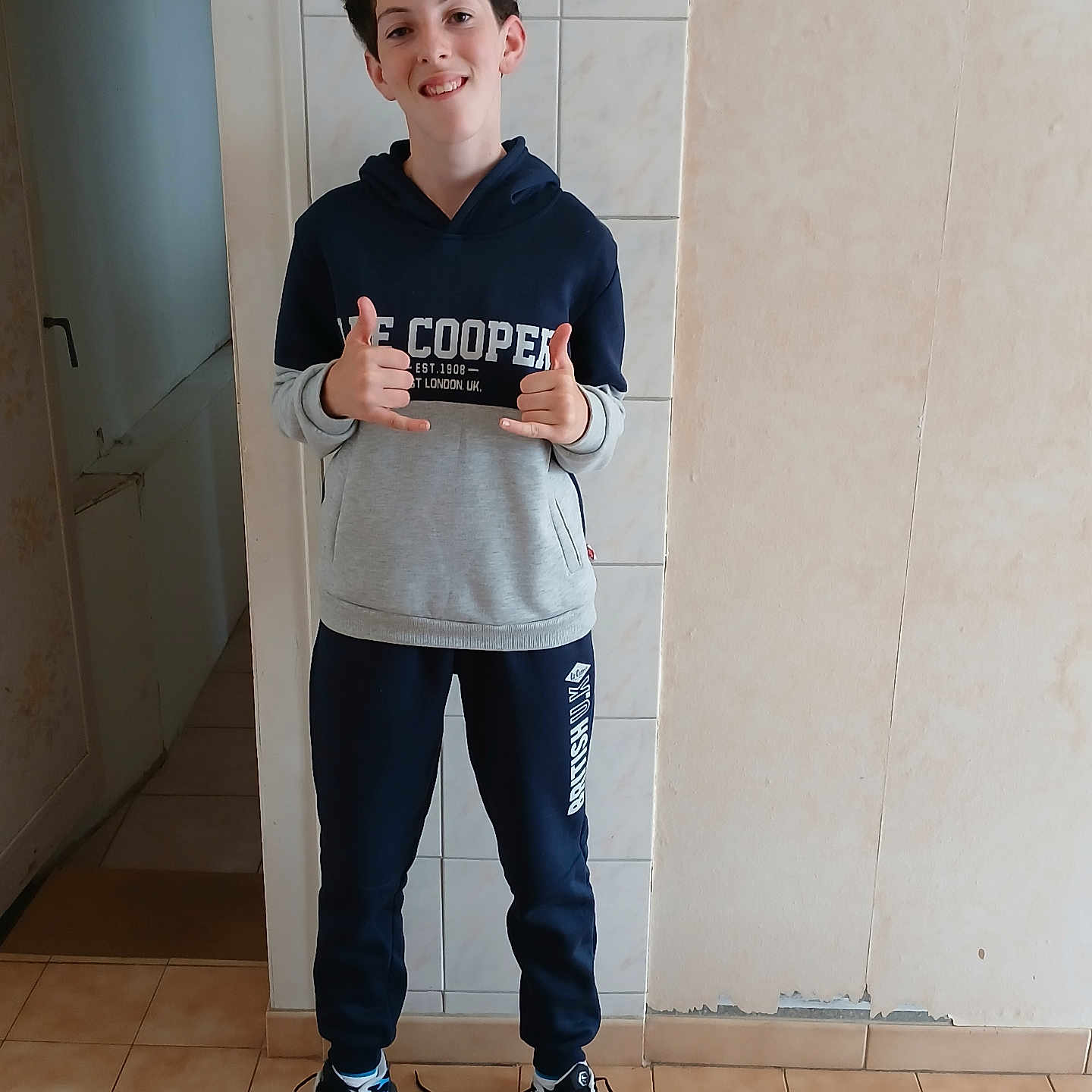 Paloma joined the competition — help win amazing prizes! boy, casual_clothing, child, curly_hair, full_body, gesture, happy, home, hoodie, indoor, person, portrait, smile, sneakers, standing, sweatpants, tile_floor, untied_shoes, wall, young