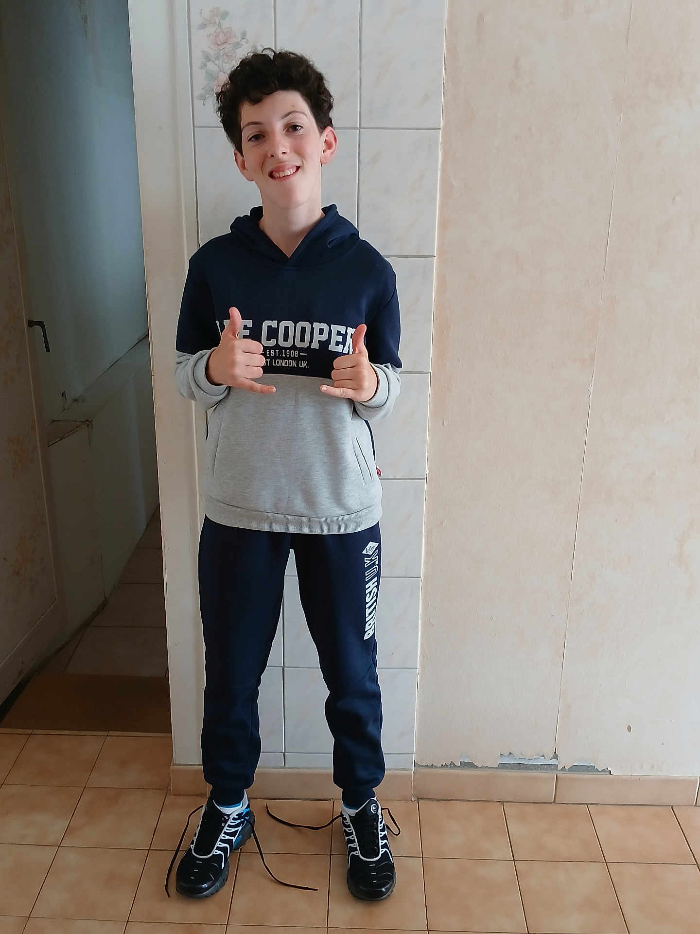 Paloma joined the competition — help win amazing prizes! boy, child, smile, indoor, casual_clothing, hoodie, sweatpants, sneakers, untied_shoes, standing, tile_floor, wall, gesture, happy, young, person, curly_hair, full_body, portrait, home