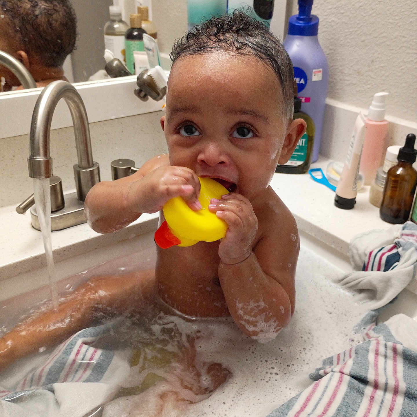 Mossiah is registered to the contest to win money with this photo: baby, bath, bathtub, bubbles, child, curly_hair, face, foam, faucet, mirror, plastic_toy, rubber_duck, sink, soap, striped_towel, water, wet_skin, young_child, bathroom, personal_care