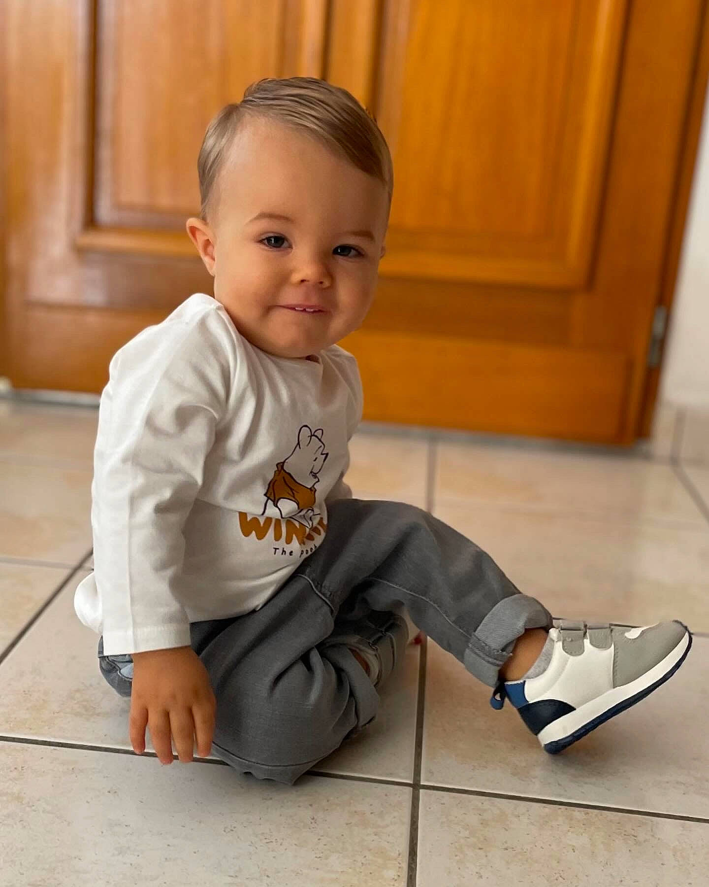 Milo participe au concours pour gagner de l'argent avec cette photo : baby, baby_toddler_clothing, clothing, comfort, door, floor, flooring, foot, fun, happy, hardwood, human_leg, knee, person, sitting, skin, sleeve, smile, toddler, walking_shoe