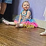 toddler, child, ballet_shoes, tutu, colorful_clothing, wooden_floor, lavender_wall, sitting, happy, smiling, person, indoor, flooring, footwear, adult_legs, dancer, dance_class, beaded_necklace, young_child, casual