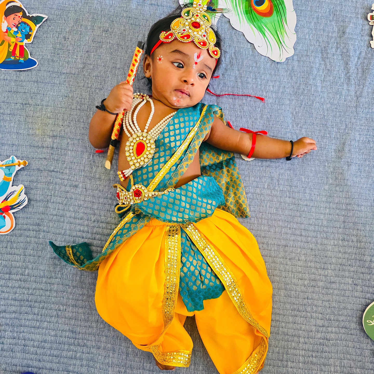 Revansh Reddy is registered to the contest to win money with this photo: baby, blue_blanket, celebration, child, colorful, costume, cute, decorations, face_paint, headband, indian_culture, indoors, infant, jewelry, lying_down, peacock_feather, stick, toy, traditional_clothing, yellow_pants