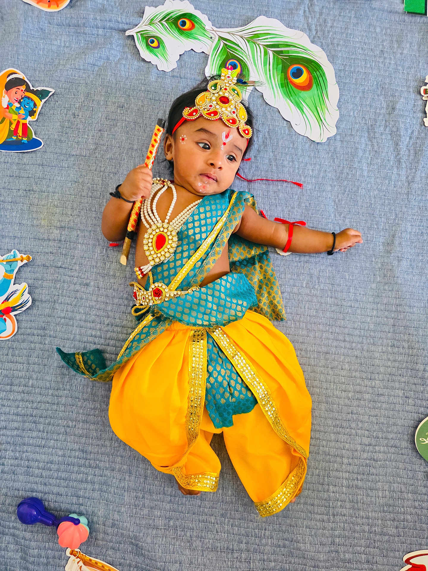 Revansh Reddy is registered to the contest to win money with this photo: baby, child, costume, traditional_clothing, peacock_feather, jewelry, blue_blanket, yellow_pants, toy, stick, cute, indian_culture, face_paint, headband, lying_down, colorful, indoors, decorations, celebration, infant
