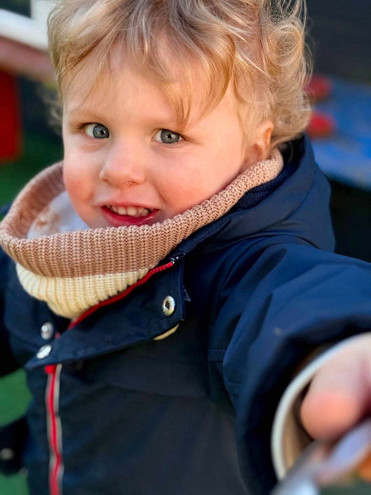 Nino a rejoint le concours — aidez-le/la à gagner de superbes lots ! child, toddler, portrait, close_up, curly_hair, blue_eyes, smile, rosy_cheeks, scarf, coat, jacket, navy, outdoor, playground, face, hand, zipper, candid, winter_clothing, young_kid