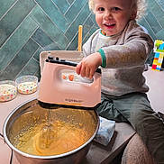 Nino a rejoint le concours — aidez-le/la à gagner de superbes lots ! child, toddler, smile, hand_mixer, mixing_bowl, batter, kitchen, countertop, teal_tiles, curly_hair, sitting, joyful, appliance, lego_blocks, starry_bowls, sweatshirt, pants, butter_packet, metal_bowl, happy_face