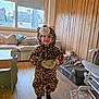 Nino participe au concours pour gagner de l'argent avec cette photo : child, leopard_costume, hood, bowl, smile, living_room, sofa, window, wood_paneling, flooring, toy, playful, socks, table, pet_bed, stuffed_animal, chair, curtain, indoor, portrait