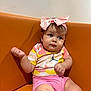 baby, child, infant, pink_shorts, colorful_shirt, headband, bow, orange_couch, sitting, indoor, cute, curious, chubby_cheeks, portrait, clothing, skin, person, young, expression, casual