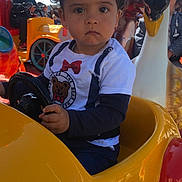 Santino a rejoint le concours — aidez-le/la à gagner de superbes lots ! toddler, child, toy_car, steering_wheel, outdoor, sunlight, carnival, ride, yellow, red, blue_sky, person, curly_hair, serious_expression, clothing, sunglasses, background, decorations, fun, playground