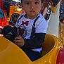 toddler, child, toy_car, steering_wheel, outdoor, sunlight, carnival, ride, yellow, red, blue_sky, person, curly_hair, serious_expression, clothing, sunglasses, background, decorations, fun, playground