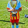 child, trophy, soccer, sports, outdoor, grass, field, sunny, blue_sky, uniform, red_shorts, blue_socks, cleats, happy, smiling, standing, celebration, youth, competition, team