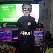 Christopher is registered to the contest to win money with this photo: child, costume, swat, sunglasses, badge, indoor, television, furniture, dog, wood_floor, fan, decor, table, blanket, person, black_clothing, smile, living_room, toy_horses, household