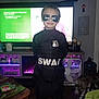 child, costume, swat, sunglasses, badge, indoor, television, furniture, dog, wood_floor, fan, decor, table, blanket, person, black_clothing, smile, living_room, toy_horses, household