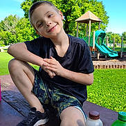 Dallas is registered to the contest to win money with this photo: child, boy, smiling, playground, slide, park, tree, grass, outdoor, summer, casual_clothing, short_hair, sneakers, picnic_table, bottle, phone, greenery, daylight, recreation, happy