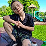 bottle, boy, casual_clothing, child, daylight, grass, greenery, happy, outdoor, park, phone, picnic_table, playground, recreation, short_hair, slide, smiling, sneakers, summer, tree