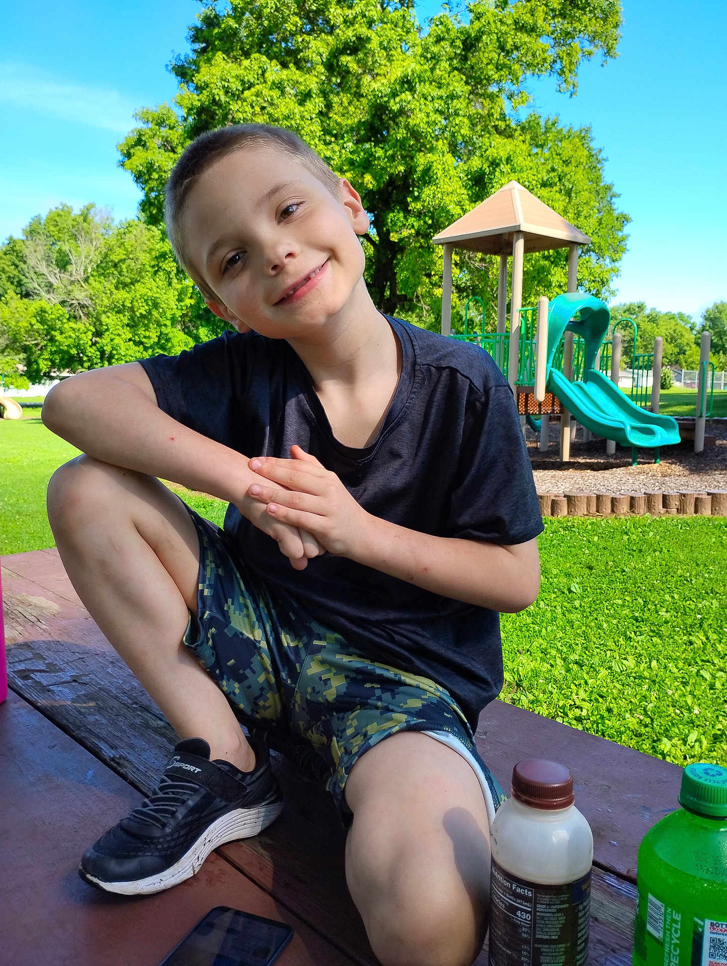 Dallas is registered to the contest to win money with this photo: child, boy, smiling, playground, slide, park, tree, grass, outdoor, summer, casual_clothing, short_hair, sneakers, picnic_table, bottle, phone, greenery, daylight, recreation, happy