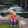 child, girl, blonde_hair, pink_shirt, jeans, pink_shoes, concrete, grass, green_car, green_suv, carport, outdoor, plants, smiling, posing, casual, daylight, nature, playful, vehicle