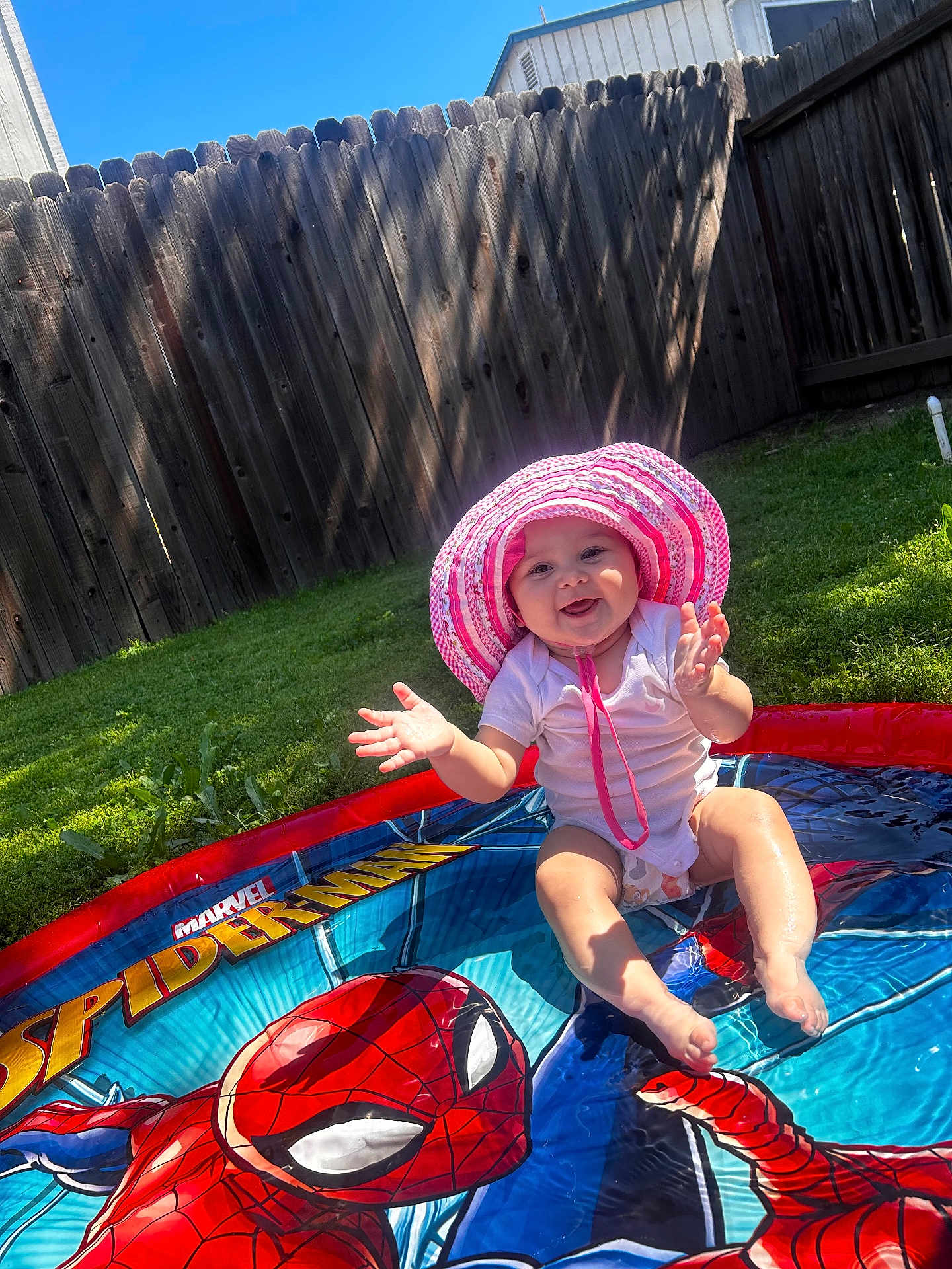 Nyleia is registered to the contest to win money with this photo: baby, child, pool, water, sunhat, pink, white_onesie, spiderman, superhero, outdoor, grass, fence, sunlight, shadow, happy, splashing, summer, play, smile, fun