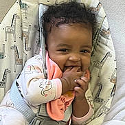 Harmony is registered to the contest to win money with this photo: baby, child, curly_hair, smiling, onesie, bib, hand_in_mouth, baby_seat, giraffe_pattern, seatbelt, indoor, cute, infant, happy, playful, person, young_child, cozy, soft_toys, comfort
