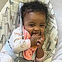 baby, child, curly_hair, smiling, onesie, bib, hand_in_mouth, baby_seat, giraffe_pattern, seatbelt, indoor, cute, infant, happy, playful, person, young_child, cozy, soft_toys, comfort