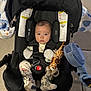baby, car_seat, infant, toy, giraffe, blue_fan, pajamas, dinosaur_pattern, face, child, sitting, clothing, safety_harness, indoor, blanket, cute, expression, head_support, footwear, person