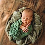 newborn, baby, sleeping, wrapped, green_cloth, basket, blanket, wooden_floor, peaceful, portrait, infant, soft_texture, cozy, resting, head, face, hands, closeup, indoor, cute