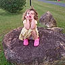 child, girl, rock, grass, tree, bush, pathway, pink_shoes, yellow_dress, curly_hair, outdoor, park, playful, nature, daylight, greenery, sitting, expression, casual_clothing, fun