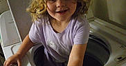 Elizabeth is registered to the contest to win money with this photo: child, washing_machine, curly_hair, laundry_room, playful, smiling, indoor, appliance, white, clothing, person, young, cute, curly, funny, home, exploring, childhood, happy, adventure
