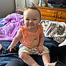 baby, child, smiling, sitting, remote_control, bed, blanket, orange_top, patterned_shorts, hair_spikes, indoors, dresser, furniture, happy, cute, person, skin, foot, hand, room