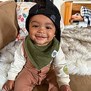 Creed is registered to the contest to win money with this photo: baby, child, infant, smiling, portrait, indoors, seated, cap, backward_cap, bib, overalls, long_sleeves, clothing, blanket, furry_blanket, couch, living_room, furniture, happy, close_up