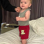 Ryker is registered to the contest to win money with this photo: air_conditioner, apparel, baby, bed, blanket, child, clothing, curtains, cute, furniture, hand, indoor, person, shorts, skin, smiling, standing, support, teddy_bear, toddler