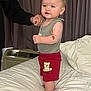 air_conditioner, apparel, baby, bed, blanket, child, clothing, curtains, cute, furniture, hand, indoor, person, shorts, skin, smiling, standing, support, teddy_bear, toddler