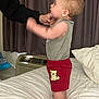 adult_hand, air_conditioner, baby, bear_patch, bed, blanket, carpet, child, clothing, curtains, cute, furniture, gray_shirt, indoor, person, pillow, red_shorts, standing, support, toddler
