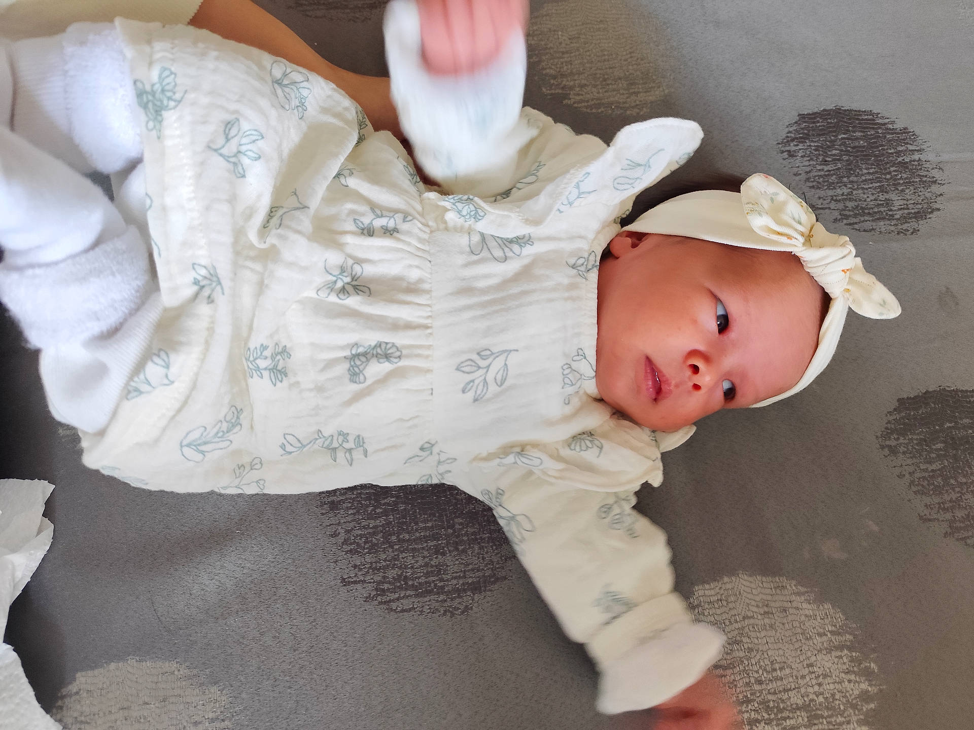 arm_motion, baby, child, curious_expression, cute, face, grey_background, headband, headwear, indoors, infant, lying_down, newborn, patterned_outfit, person, resting, sleepwear, small_hands, soft_fabric, white_clothing